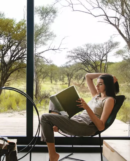 Woman reading indoors beside a large aluminium window overlooking natural scenery.